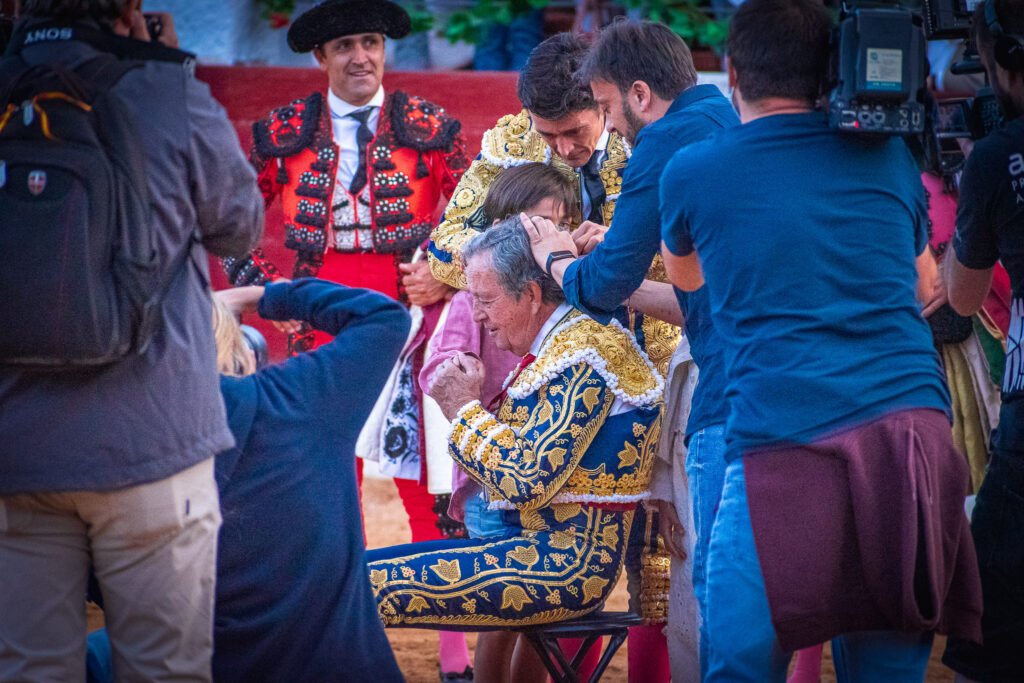 Momento emotivo del corte de coleta de un torero veterano vestido de azul y oro rodeado de compañeros y fotógrafos.